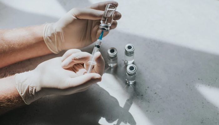 1 de 1 Uma mão com luvas de látex segura um frasco com líquido transparente e uma seringa. O p...